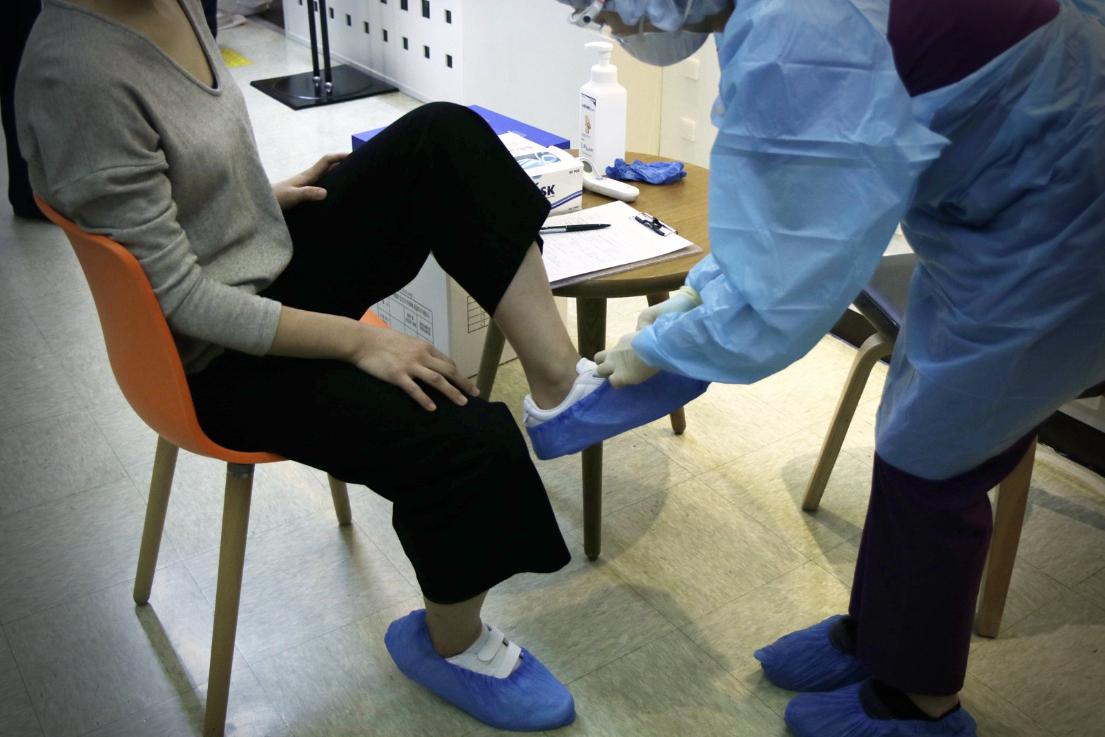 보호가운과 장갑을 착용한 직원이 몸을 굽혀 의자에 앉은 사람의 신발에 파란 일회용 덧신을 씌우고 있다. 옆 탁자에 손소독제와 마스크 상자, 비접촉 체온계와 문진표가 놓여 있다